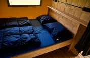 Cozy glamping accommodation with wooden bed frame, dark blue bedding, and window in the background.