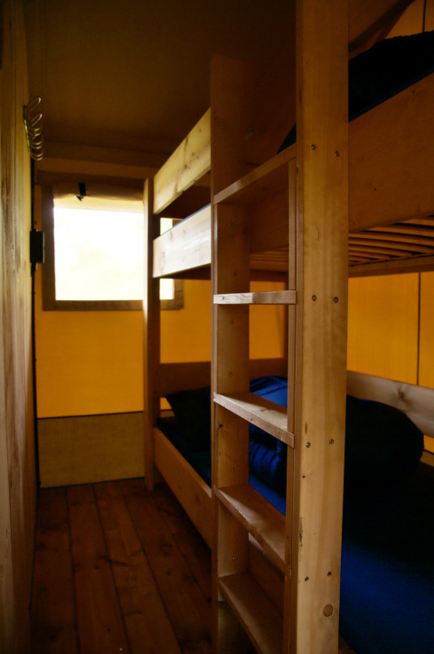 Vista interior de un alojamiento glamping con literas de madera y ropa de cama azul junto a una ventana.