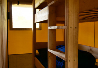 Binnenzicht van een glampingverblijf met houten stapelbedden en blauw beddengoed bij het raam.