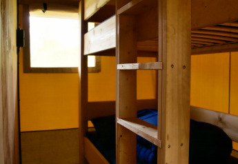 Vista interior de un alojamiento glamping con literas de madera y ropa de cama azul junto a una ventana.
