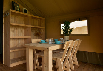 Wooden dining table and chairs inside a safari tent with shelves and plant at Camping La Dolce Vita.