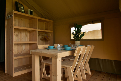 Wooden dining table and chairs inside a safari tent with shelves and plant at Camping La Dolce Vita.