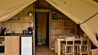 Interieur van een glampingtent met kitchenette, koelkast, houten eettafel en stoelen zichtbaar.
