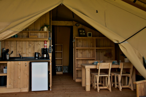 Interieur van een glampingtent met kitchenette, koelkast, houten eettafel en stoelen zichtbaar.