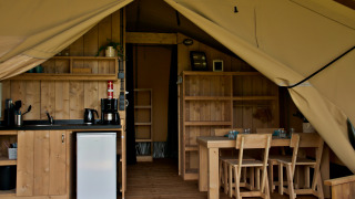 Interior of glamping tent with kitchenette, mini fridge, wooden dining table, and chairs visible.