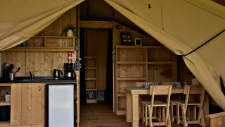 Interior of glamping tent with kitchenette, mini fridge, wooden dining table, and chairs visible.
