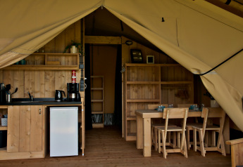 Interior of glamping tent with kitchenette, mini fridge, wooden dining table, and chairs visible.