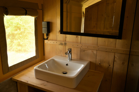 Lavabo moderno e specchio in una tenda safari, circondato da pareti di legno e luce naturale.
