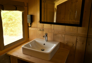 Lavabo moderne et miroir dans une tente safari, murs en bois et lumière naturelle venant de la fenêtre.