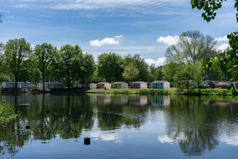 Parque vacacional con cabañas de glamping junto a un lago, rodeado de árboles y cielo azul.