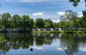Feriepark med glamping-hytter ved en sø, omgivet af grønne træer og blå himmel med skyer.
