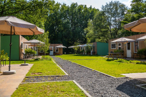 Udendørsbillede af glampingboliger i en feriepark med grønne områder, solparasoller og træhytter.