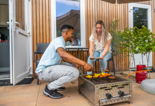 Par griller mad sammen udenfor et moderne glamping-hytte på en ferielejr, afslappet stemning.