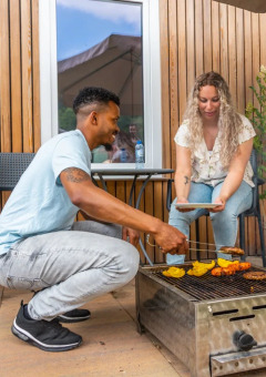 Pareja asando comida juntos afuera de una moderna cabaña glamping en un parque vacacional, ambiente relajado.
