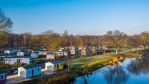 Feriepark med glampingindkvartering, små hytter og sø omgivet af træer under blå himmel.
