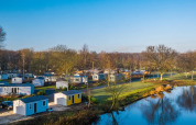 Feriepark med glampingindkvartering, små hytter og sø omgivet af træer under blå himmel.