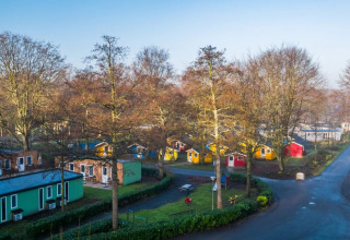 Luftfoto af ferielejr med farverige glamping-hytter omgivet af træer og små veje i naturskønt område.