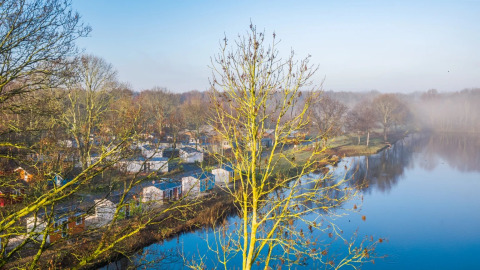 Blick auf einen Ferienpark mit Glamping-Unterkünften am See, umgeben von Bäumen im Morgennebel.