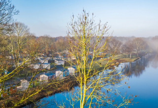 Blick auf einen Ferienpark mit Glamping-Unterkünften am See, umgeben von Bäumen im Morgennebel.