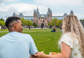 Twee mensen picknicken op het gras voor het Rijksmuseum in Amsterdam op een zonnige dag.