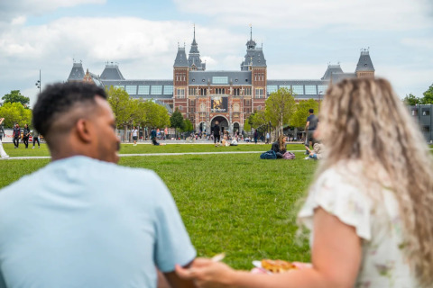 Twee mensen picknicken op het gras voor het Rijksmuseum in Amsterdam op een zonnige dag.