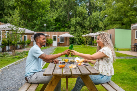 Pareja disfruta comida al aire libre y brinda en mesa de picnic en parque vacacional con glamping detrás.