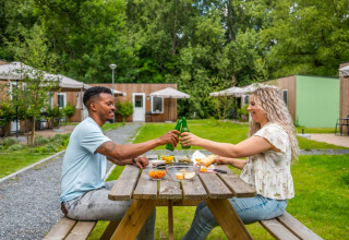 Pareja disfruta comida al aire libre y brinda en mesa de picnic en parque vacacional con glamping detrás.