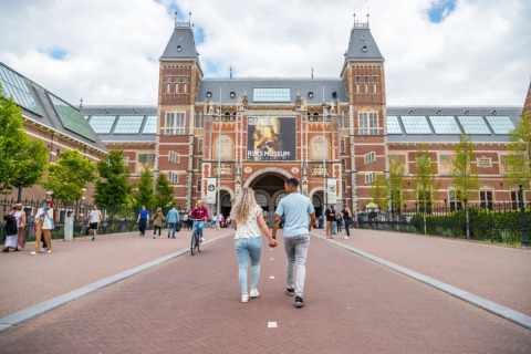 A couple walks hand in hand towards the Rijksmuseum in Amsterdam, surrounded by other visitors outdoors.
