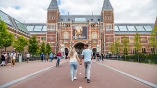 Una pareja camina de la mano hacia el Rijksmuseum en Ámsterdam, rodeada de otros visitantes y árboles.