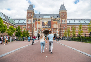 Et par går hånd i hånd mod Rijksmuseum i Amsterdam, omgivet af andre besøgende på en solrig dag.