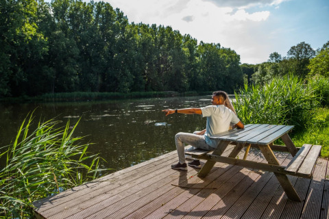 Couple on a bench by a lake in a glamping park, enjoying the view of the water and lush green forest.