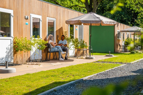 Een stel ontspant voor moderne glamping-accommodaties met parasol in een zonnig, groen vakantiepark.