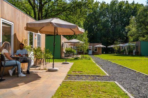 Dos personas desayunan frente a cabañas de madera con sombrillas en un glamping de un parque vacacional.