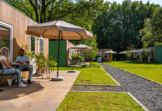 Deux personnes prennent le petit-déjeuner devant des cabanes en bois avec parasols dans un parc de glamping.