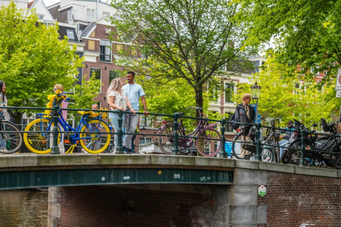Mensen lopen en staan op een brug in de stad met kleurrijke fietsen en groene bomen op de achtergrond.