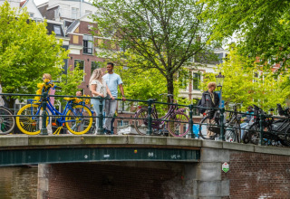 People walking and standing on a city bridge with colorful bicycles and green trees in the background.