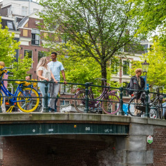 Personas caminando y de pie sobre un puente urbano con bicicletas coloridas y árboles verdes detrás.