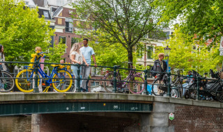 Personas caminando y de pie sobre un puente urbano con bicicletas coloridas y árboles verdes detrás.