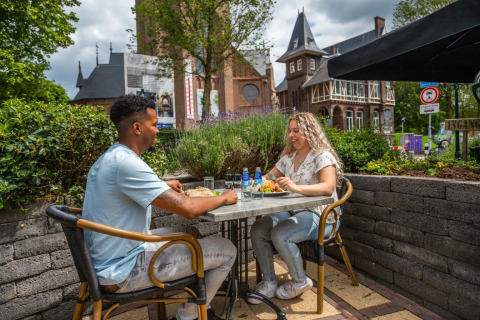 Twee personen lunchen buiten bij een vakantiepark met glamping, omgeven door groen en ontspannen sfeer.