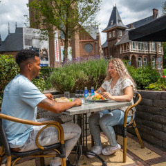 Dos personas almuerzan al aire libre en un parque vacacional con glamping, rodeados de naturaleza y jardines.