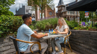 Dos personas almuerzan al aire libre en un parque vacacional con glamping, rodeados de naturaleza y jardines.