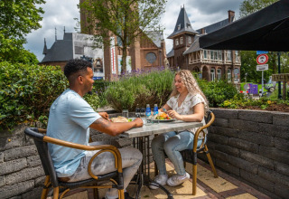 To personer nyder en udendørs frokost på en café i en feriepark med glamping, omgivet af grønne omgivelser.