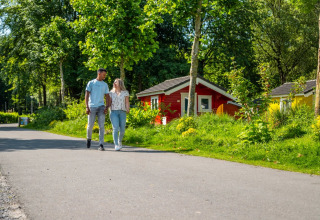 Et par går hånd i hånd foran farverige glamping-hytter i en frodig feriepark omgivet af natur.