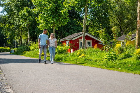 Et par går hånd i hånd foran farverige glamping-hytter i en frodig feriepark omgivet af natur.