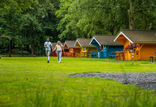 Par går hånd i hånd foran farverige glamping-hytter i en feriepark omgivet af grønne træer og græs.