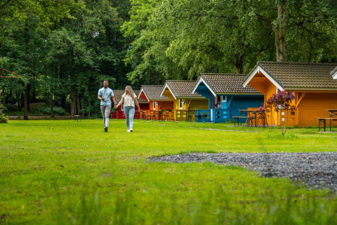 Pareja caminando de la mano frente a coloridas cabañas glamping en un parque vacacional rodeado de árboles y césped.