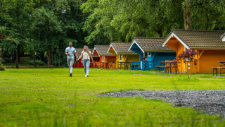 Pareja caminando de la mano frente a coloridas cabañas glamping en un parque vacacional rodeado de árboles y césped.