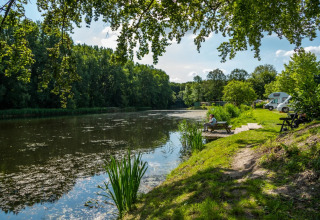 Feriepark ved sø med træer, autocamper og gæst på bænk. Perfekt til glamping og naturferie.