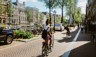 Woman cycling along a scenic canal lined with classic buildings and trees on a sunny day in the city.