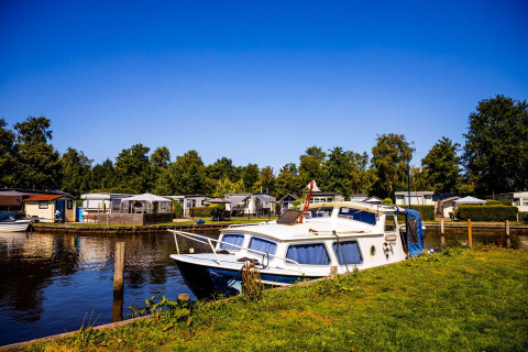 A boat docked by the shore of a lake at a holiday park in Friesland, Netherlands, with cabins and greenery.